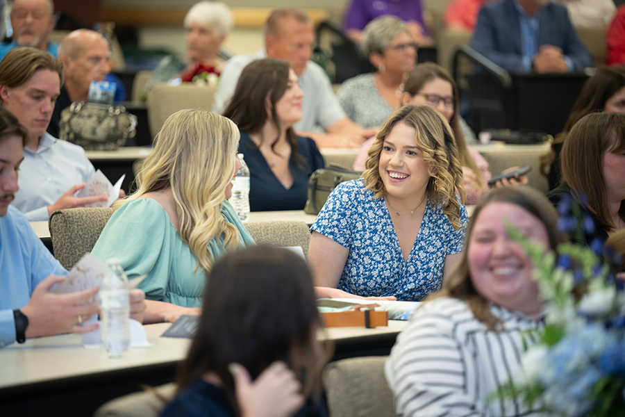 A smiling, blonde female student, wearing a white dress with black line patterns on it, stands to the left of a smiling female faculty member with dark hair who is wearing a white and lavender dress with a grey and lavender scarf. They both look forward while posing for a photo in a classroom setting with the photographer visible on the right side of the frame and out of focus.  